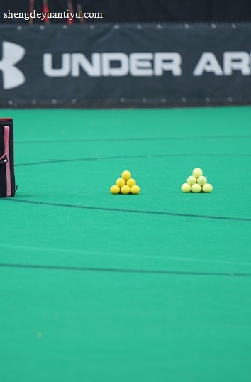 A sports field features a black and pink sports bag, two pyramids of yellow balls, and a portion of a person in dark athletic gear. The background displays a banner with the Under Armour logo.