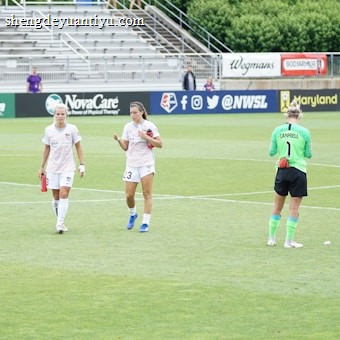 Three athletes are on a soccer field; two are wearing white sports uniforms while one is in a green goalkeeper outfit. They appear to be walking across the field, with a background of empty stands and various advertisements.