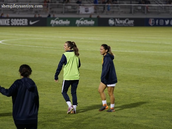Two people are on a soccer field, wearing sports attire. One person is wearing a green vest, and the other is in a blue jacket and white shorts. The setting appears to be during a sports event, with adverts in the background and a partially filled stadium.