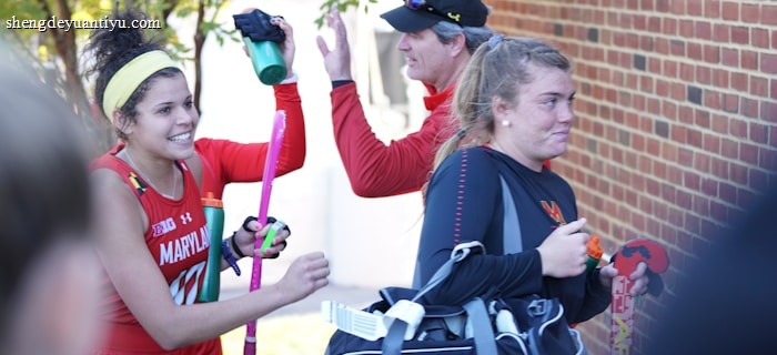 A group of athletes in sports attire appears to be in a joyful mood. One person is holding a field hockey stick and wearing a headband, while another carries a sports bag. They are gathered near a brick wall with trees in the background, sharing a light moment, likely after a sports event.