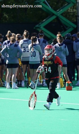 A sports team gathers on a field with a coach, all dressed in athletic gear, while one player wearing a red helmet and jersey walks away from the group carrying a lacrosse stick. The field is green, and there are sports equipment and water bottles visible.