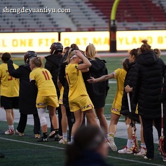 A group of athletes wearing yellow sports uniforms are standing on a sports field. Some are holding lacrosse sticks, and they appear to be interacting casually. Several people are dressed in black coats. In the background, there is a bright sign with text and parts of a stadium are visible.