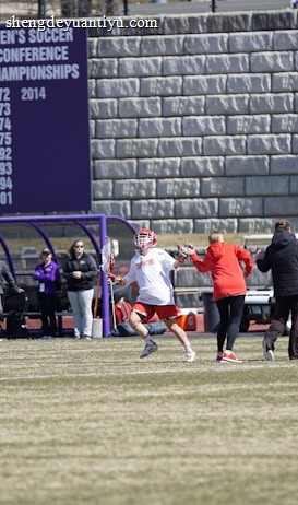 A sports team is practicing or playing on a grassy field in front of a large stone wall with banners displaying men's soccer achievements. People are wearing sports attire, and some are engaged in conversation or coaching.