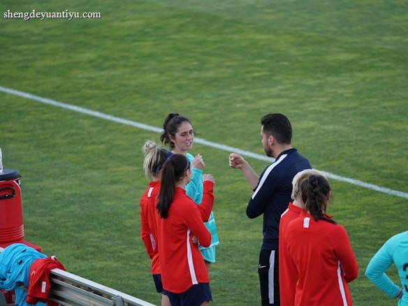 A group of people wearing sports attire are gathered on a grassy field. Some individuals are dressed in red jackets, while others are in turquoise and navy blue. A coach appears to be giving instructions. Sports equipment, such as water bottles and a bench with towels, can be seen nearby.