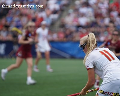 A sporting event with athletes on a grass field, one in a white jersey with the number 11 in the foreground, holding a lacrosse stick. Players in the background wear maroon uniforms. Spectators fill the stands creating a lively atmosphere.