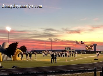 A sports field with a football game unfolding under a vibrant sunset sky, featuring a large inflatable mascot and a scoreboard displaying the score. Players and coaches are visible on the green field, surrounded by a track and some flags waving in the breeze.